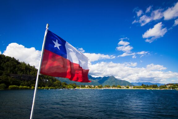 Chilean flag waving in the foreground with a clear blue sky and a scenic lake and mountains in the background.