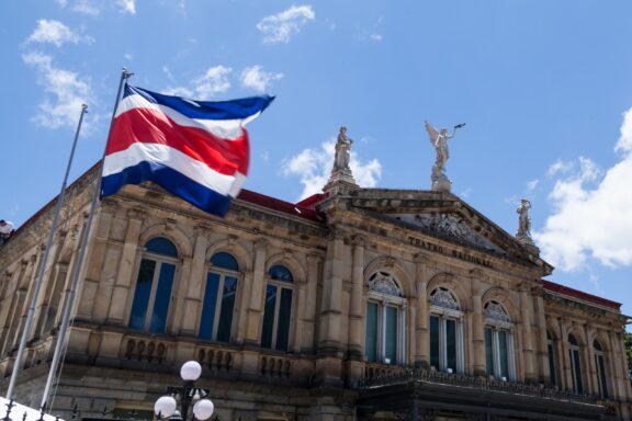 Costa Rican flag waving in front of a classical building with statues and clear blue sky.