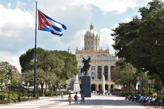 Cuban flag waving in front of a historic building with a dome in Havana, with people walking in the foreground.