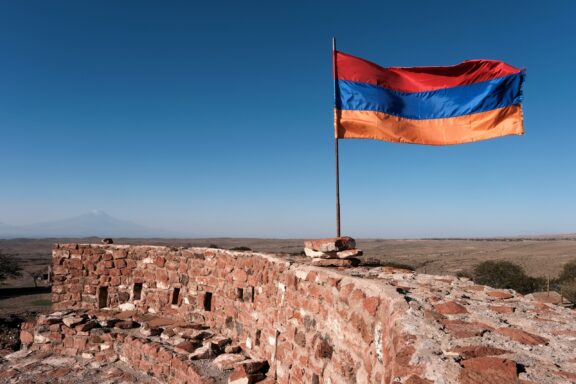 Armenian flag waving above a stone structure with a clear blue sky in the background.