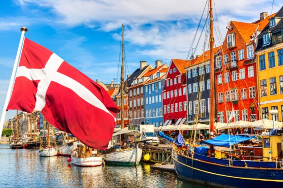 Danish flag waving in front of colorful buildings at Nyhavn port, Copenhagen, with boats docked in the water.