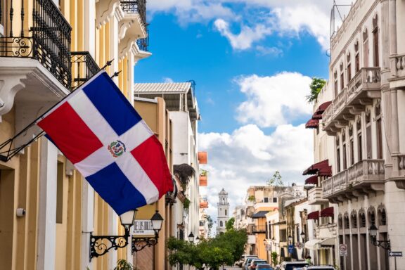 Dominican Republic flag waving in front of a street with colonial buildings under a partly cloudy sky.