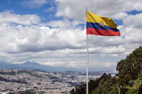 Ecuadorian flag waving against a backdrop of cloudy sky with a panoramic view of Quito city below.