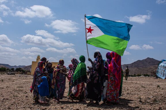 Group of people in traditional clothing gathered around the flag of Djibouti with a landscape in the background.