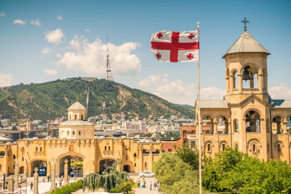 Flag of Georgia flying above Tbilisi with traditional architecture and hills in the background.