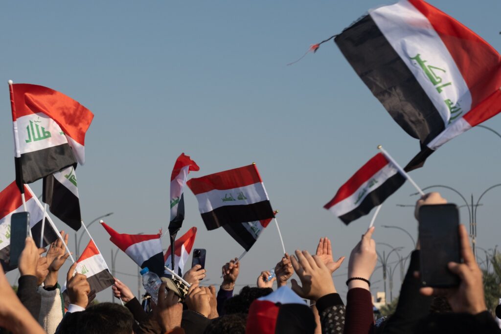 Crowd of people waving Iraqi flags against a clear sky.