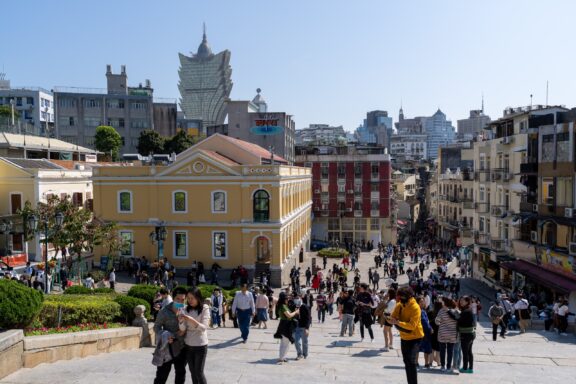 A crowd of people stand outside on a sunny day in Macau, China.