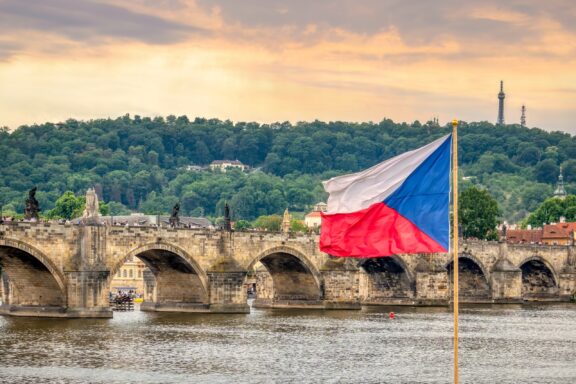 Czech flag waving in front of the Charles Bridge and Vltava River at sunset in Prague.