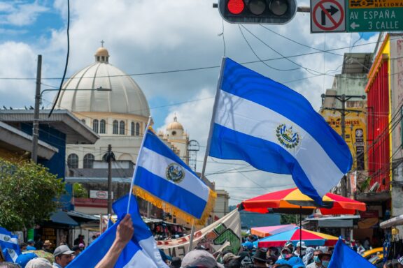 Flags of El Salvador being waved in a street with a crowd and a dome structure in the background.