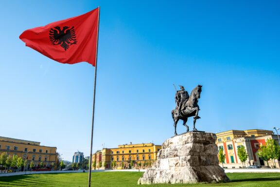 Albanian flag waving in front of the Skanderbeg statue at Skanderbeg Square with clear blue sky.