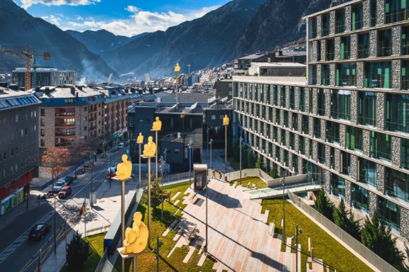 Sculptures of men sitting on poles stand in the city of Andorra la Vella with the Pyrenees in the background.