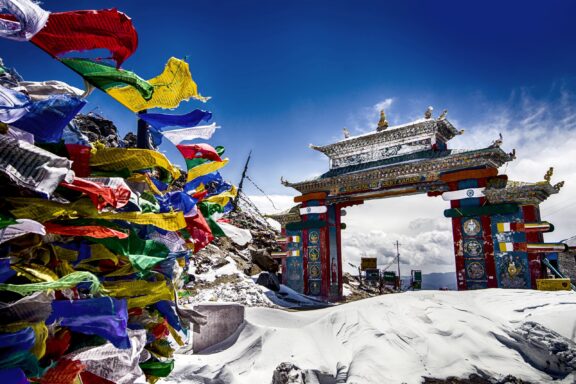 Prayer flags fly in the wind next to Buddhist architecture and snow-covered ground in Tawang.