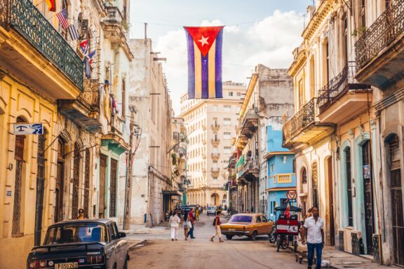 People and cars occupy a street with a Cuban flag in Havana, Cuba.