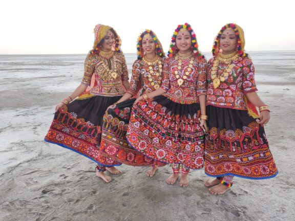 Four female dancers wearing ornate, colorful clothing stand on a great salt marsh.