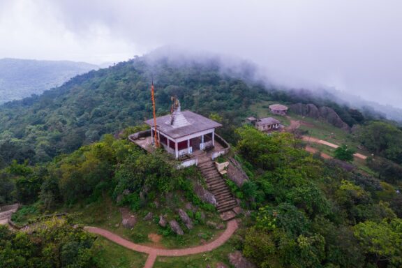 An aerial view of a small temple atop a green hill with low-lying clouds surrounding it.