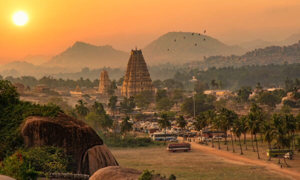 A temple stands tall in the distance in Hampi as birds fly across the sky and the sun sets.