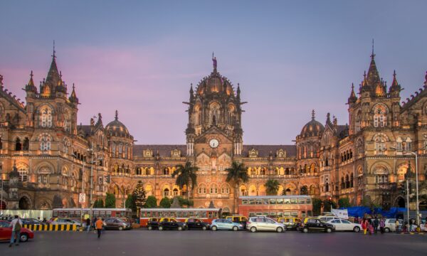 The railway building is illuminated at dusk, and cars and buses line up in front.