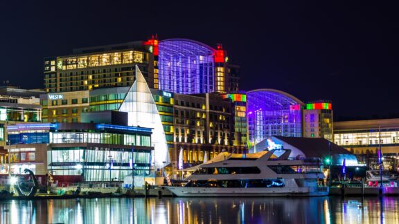 Colorful buildings and a boat reflect in the water at night in National Harbor, Maryland.