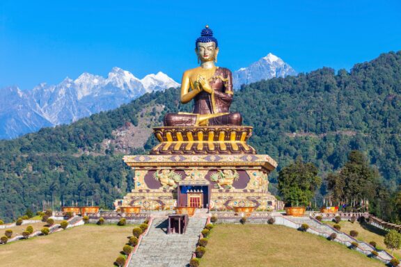 A golden statue sits atop a large stone pedestal with a mountainous backdrop in Sikkim.