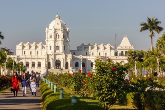 Tourists walk in front of the Ujjayanta Palace on a sunny day.