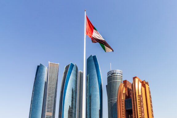 Flag of the United Arab Emirates waving in front of modern skyscrapers against a clear blue sky.