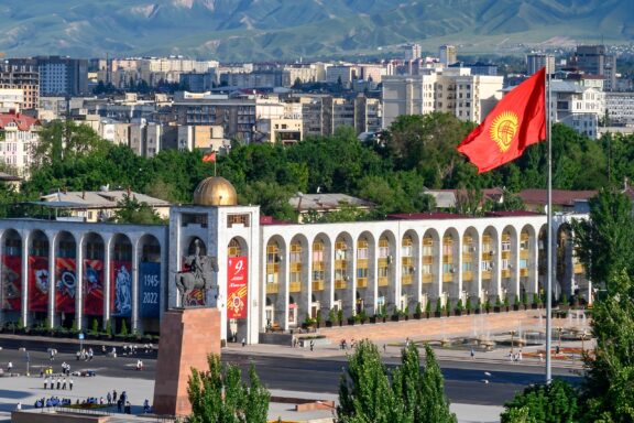 Flag of Kyrgyzstan waving above a cityscape with mountains in the background.