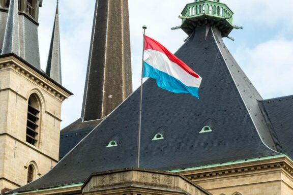 Luxembourg flag waving on top of the spire of the Notre-Dame Cathedral against a cloudy sky.