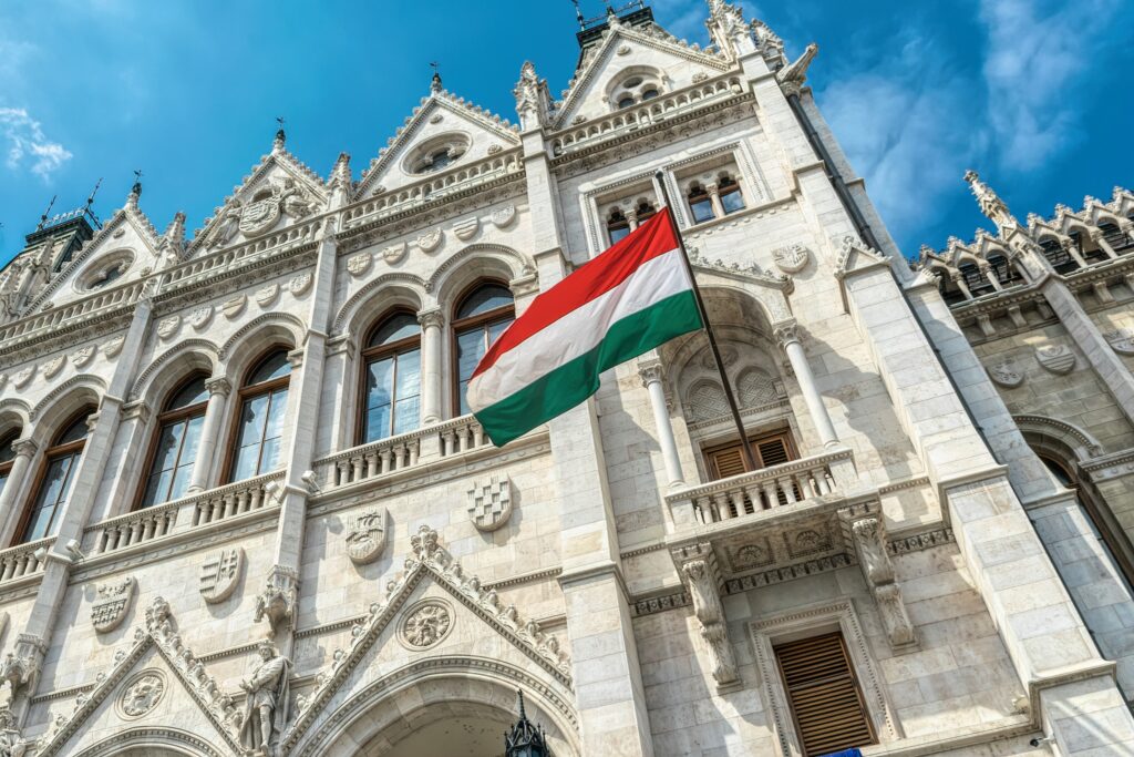 Hungarian flag waving in front of the ornate facade of the Budapest Parliament building under a clear blue sky.