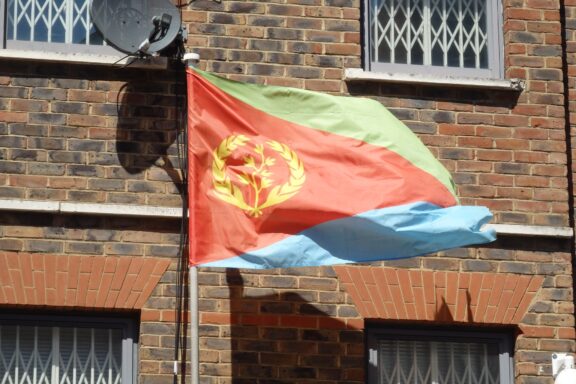 The Eritrean flag, with its green, blue, and red triangles and olive branch within a wreath centered on the red triangle, flutters against a brick building backdrop.