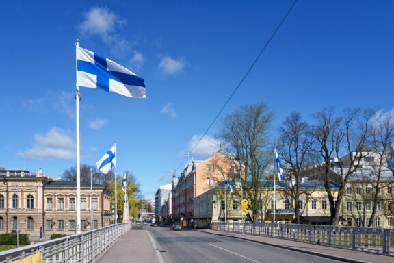 Finnish flags fluttering above a bridge with buildings and trees in the background on a clear day.