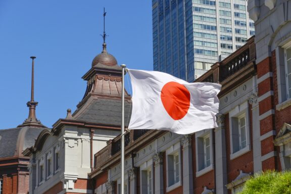 Japanese flag fluttering in front of traditional and modern buildings under a clear blue sky.