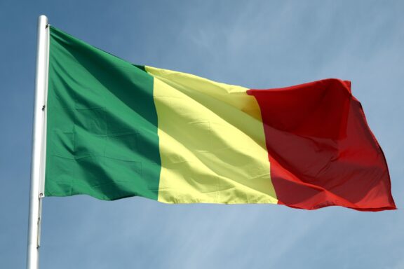 Flag of Mali with vertical stripes in green, yellow, and red fluttering against a blue sky.