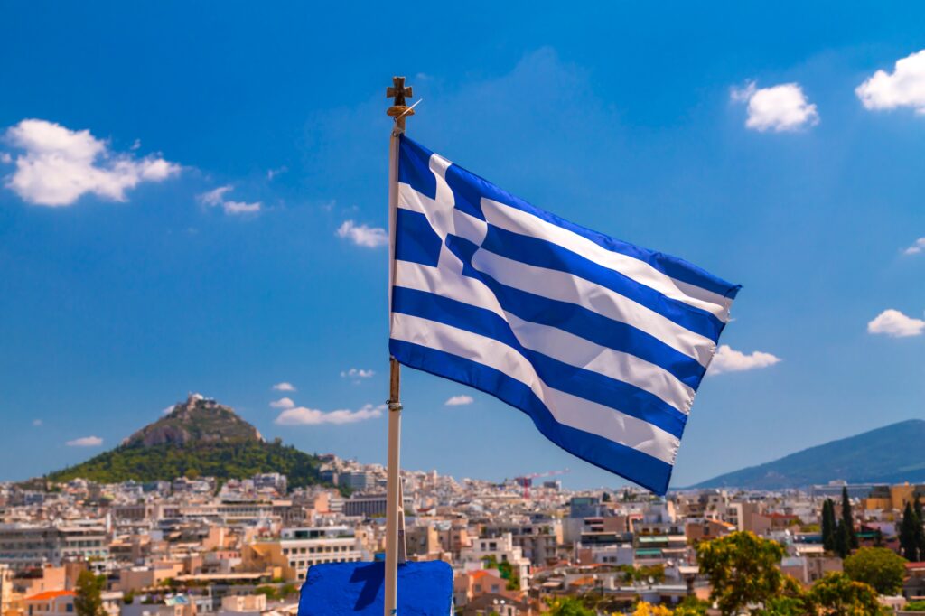 Greek flag waving with Athens cityscape and Lycabettus Hill in the background.