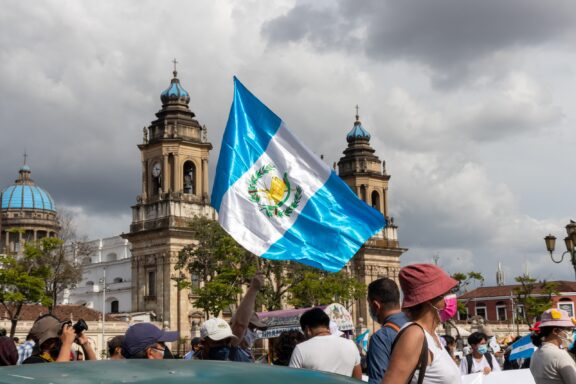 Guatemalan flag waving in front of historic buildings with cloudy sky and people in the foreground.