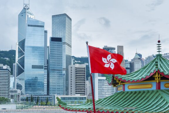 Hong Kong flag waving in front of modern skyscrapers and a traditional Chinese pavilion.