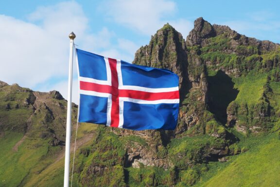 Icelandic flag waving in front of green mountains under a blue sky.