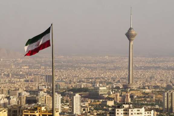 Iranian flag waving with the Tehran skyline and Milad Tower in the background.