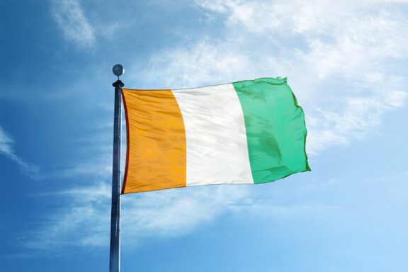 The flag of Ivory Coast, consisting of three vertical stripes in orange, white, and green, fluttering against a blue sky.