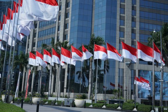 Row of Indonesian flags fluttering in front of modern office buildings under a clear blue sky.