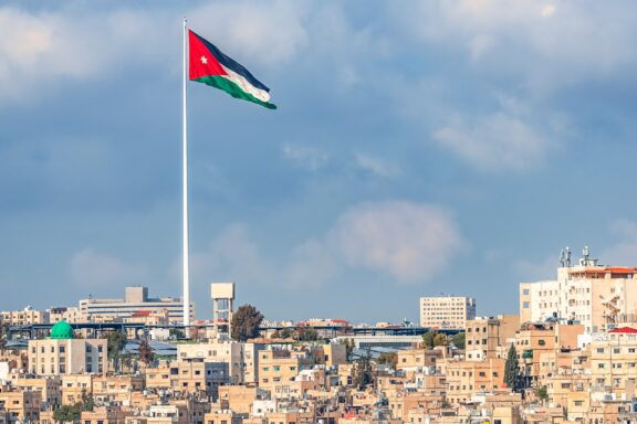 Jordanian flag flying high above the cityscape of Amman under a cloudy sky.