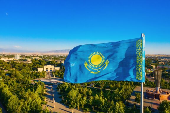 Kazakhstan flag waving against a clear blue sky above a lush green park.