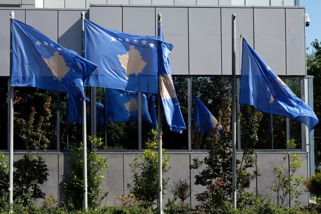 Three Kosovo flags fluttering on flagpoles in front of a building with green foliage.