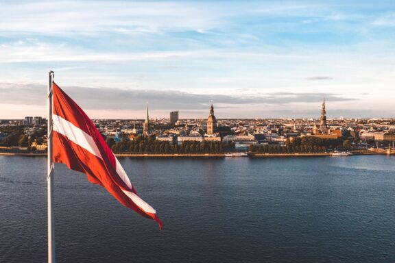 Latvian flag waving in the foreground with a panoramic view of Riga's skyline and river in the background.