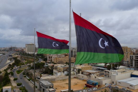 Two Libyan flags fluttering above a cityscape with buildings and roads on an overcast day.