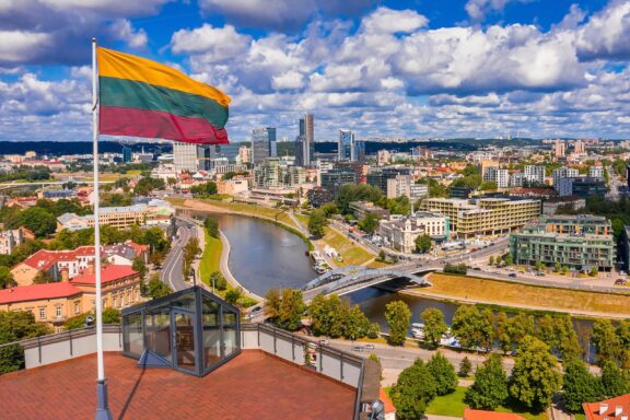 Lithuanian flag waving in the foreground with a panoramic view of Vilnius cityscape and river in the background.