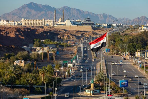 Egyptian flag waving in the foreground with the landscape of Sharm El Sheikh, including buildings and mountains, in the background.