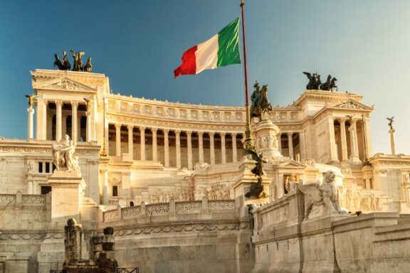 Italian flag waving in front of the Vittoriano Monument in Rome under a clear sky.