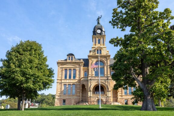 A low-angle view of the Blue Earth County Courthouse behind a tree and an American flag in Mankato, Minnesota.