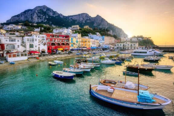 Boats sit in the water next to colorful buildings on the shore at sunset in Capri, Italy.