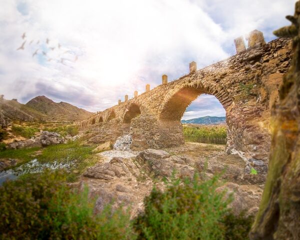 A low-angle view of a stone bridge passing over rugged terrain.